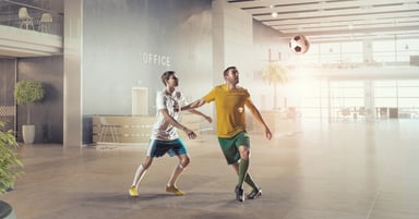 Two soccer players in athletic uniforms compete for the ball inside a modern office lobby
